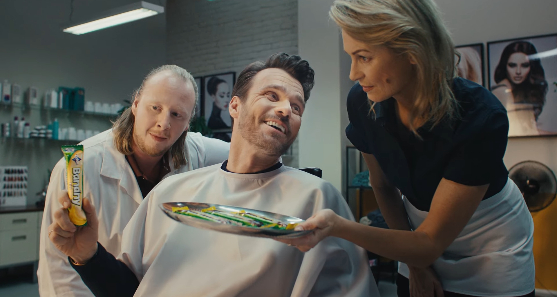 Muž sedí u kadeřníka s pláštěm, druhý muž ukazuje tyčinku Banány, žena kadeřnice předkládá nabídku produktů na vlasy.