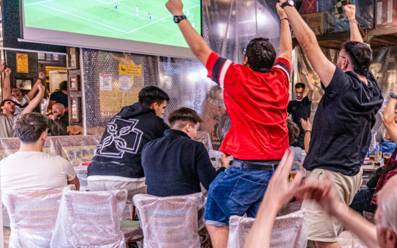 Lidé sledují fotbalový zápas na velké obrazovce v rušné hospodě, někteří nadšeně stojí a zvedají ruce.