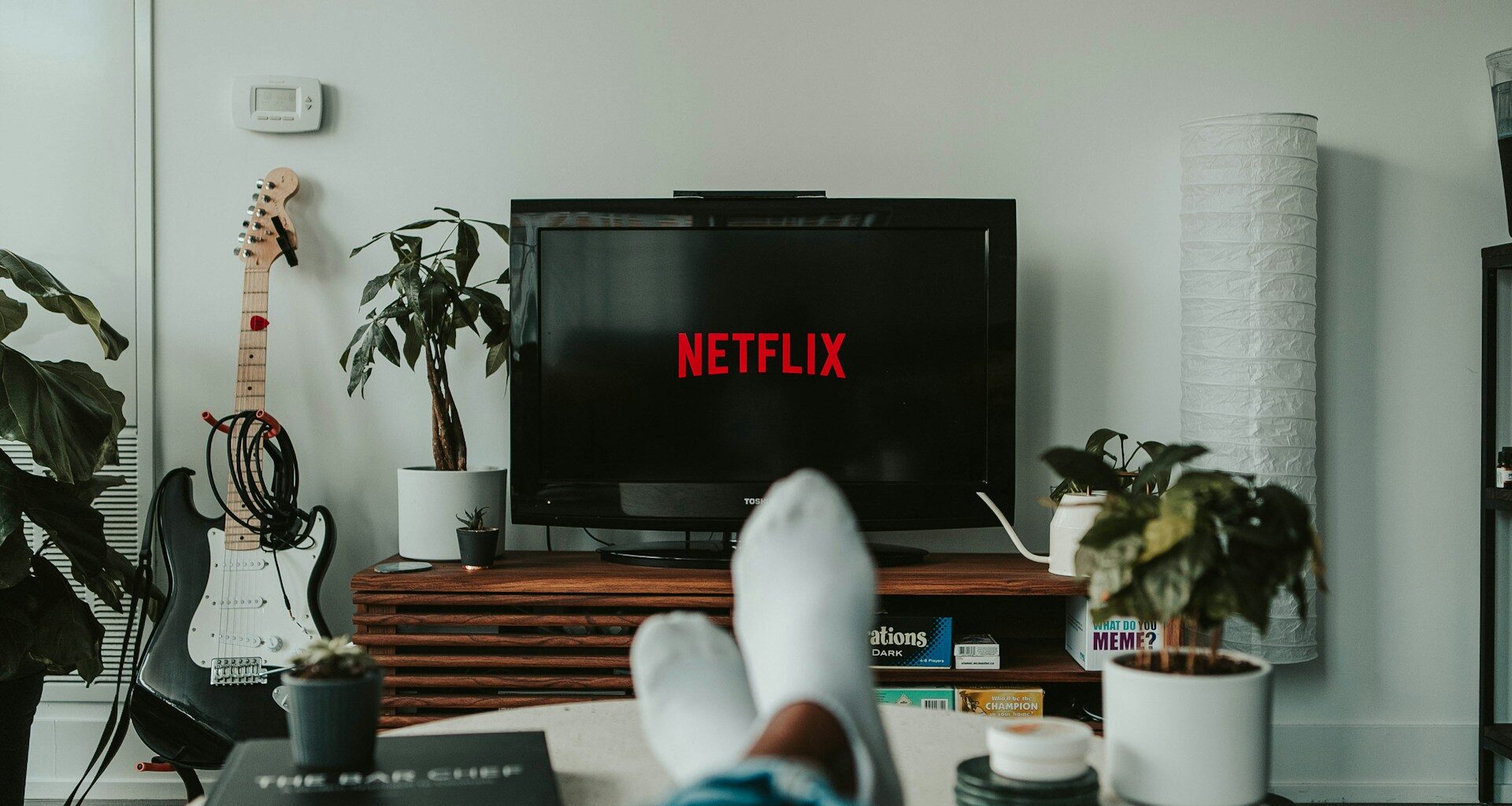 Osoba ležící na pohovce před televizí s logem Netflix; v místnosti jsou rostliny a kytara.