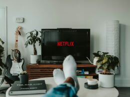 Osoba ležící na pohovce před televizí s logem Netflix; v místnosti jsou rostliny a kytara.