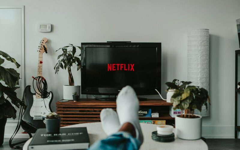 Osoba ležící na pohovce před televizí s logem Netflix; v místnosti jsou rostliny a kytara.