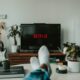 Osoba ležící na pohovce před televizí s logem Netflix; v místnosti jsou rostliny a kytara.