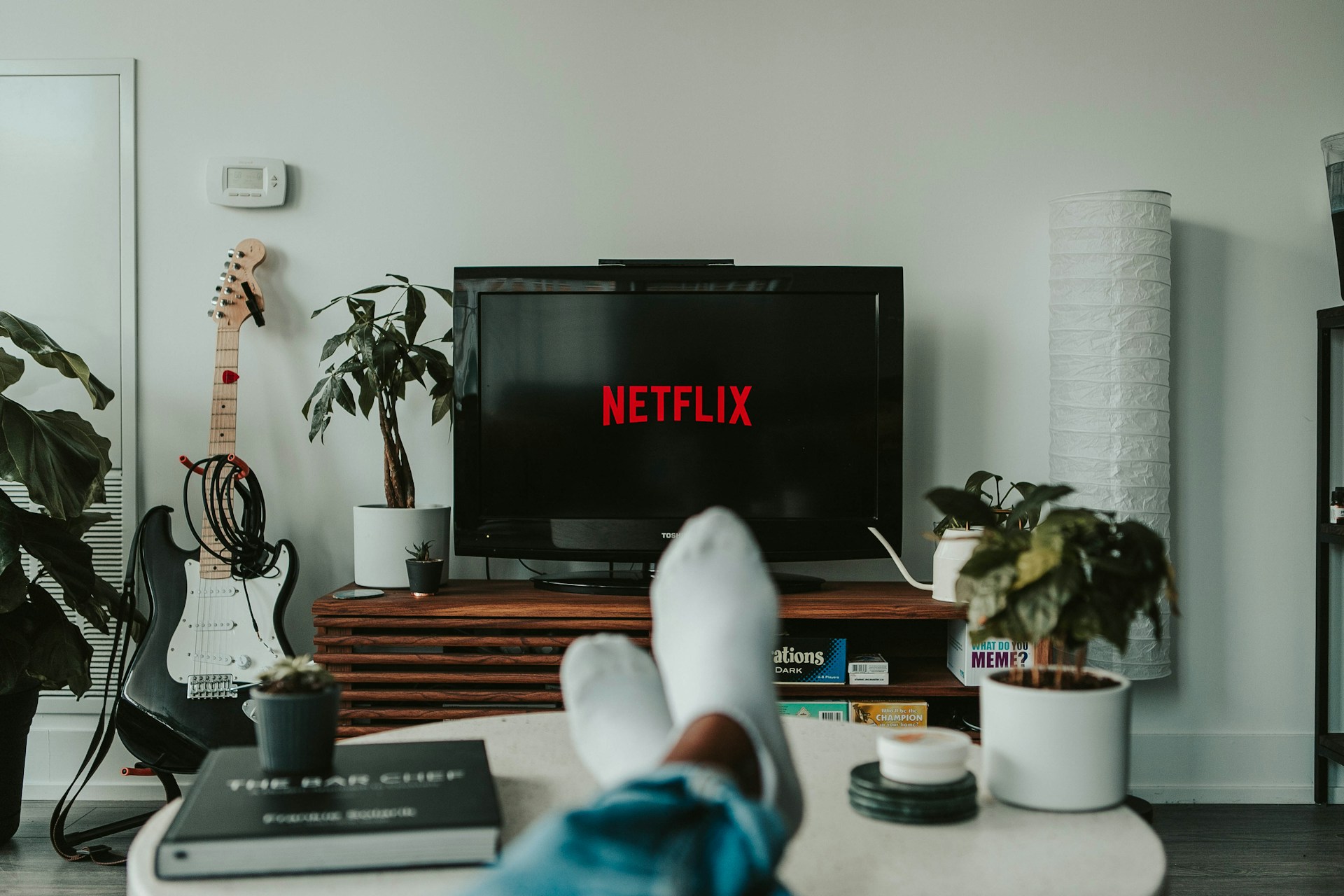 Osoba ležící na pohovce před televizí s logem Netflix; v místnosti jsou rostliny a kytara.