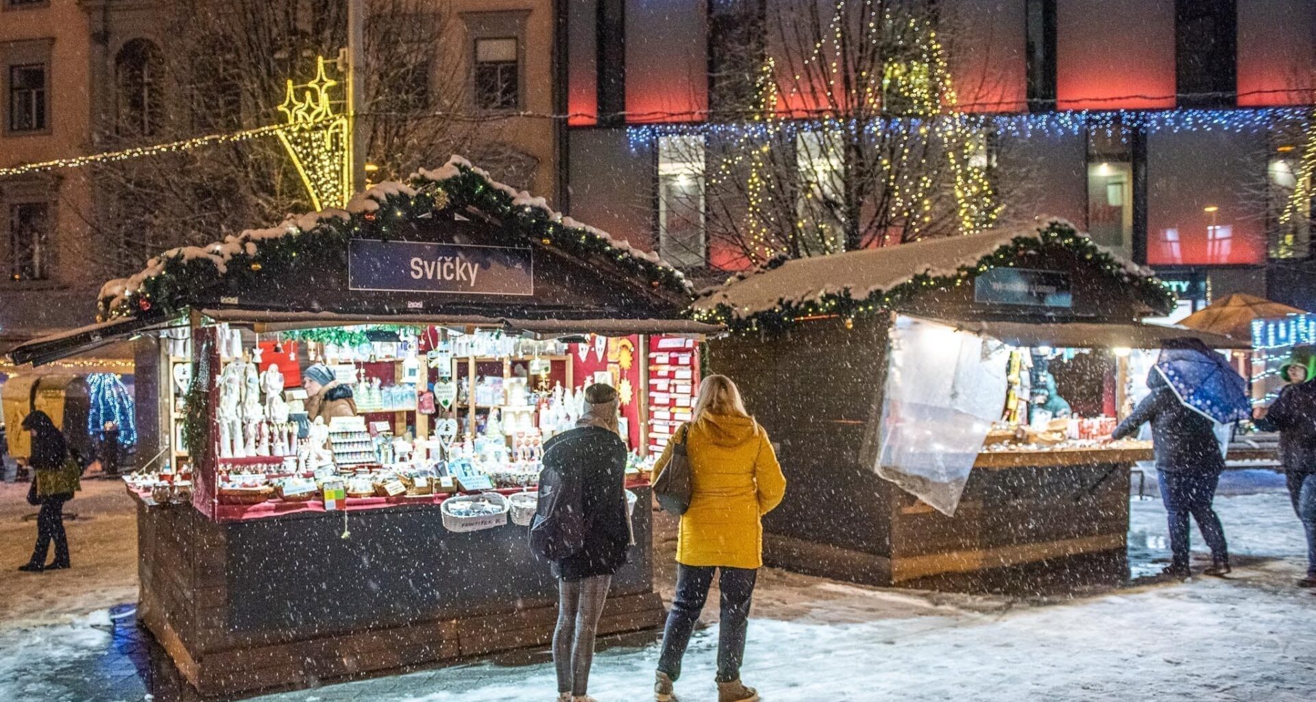 Trh s vánočními stánky, lidé nakupují svíčky, zasněžené okolí a světelná výzdoba.