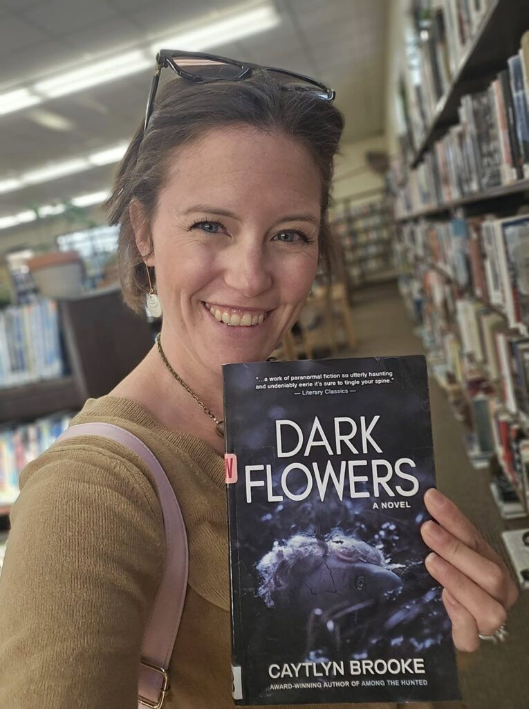 Autorka v knihovně drží knihu "Dark Flowers" od Caytlyn Brooke.