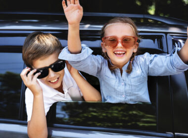 Dvě děti v autě se slunečními brýlemi, smějí se a mávají.