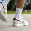 Muž v bílých teniskách Reebok s vínovými detaily a bílých ponožkách kráčí po chodníku.