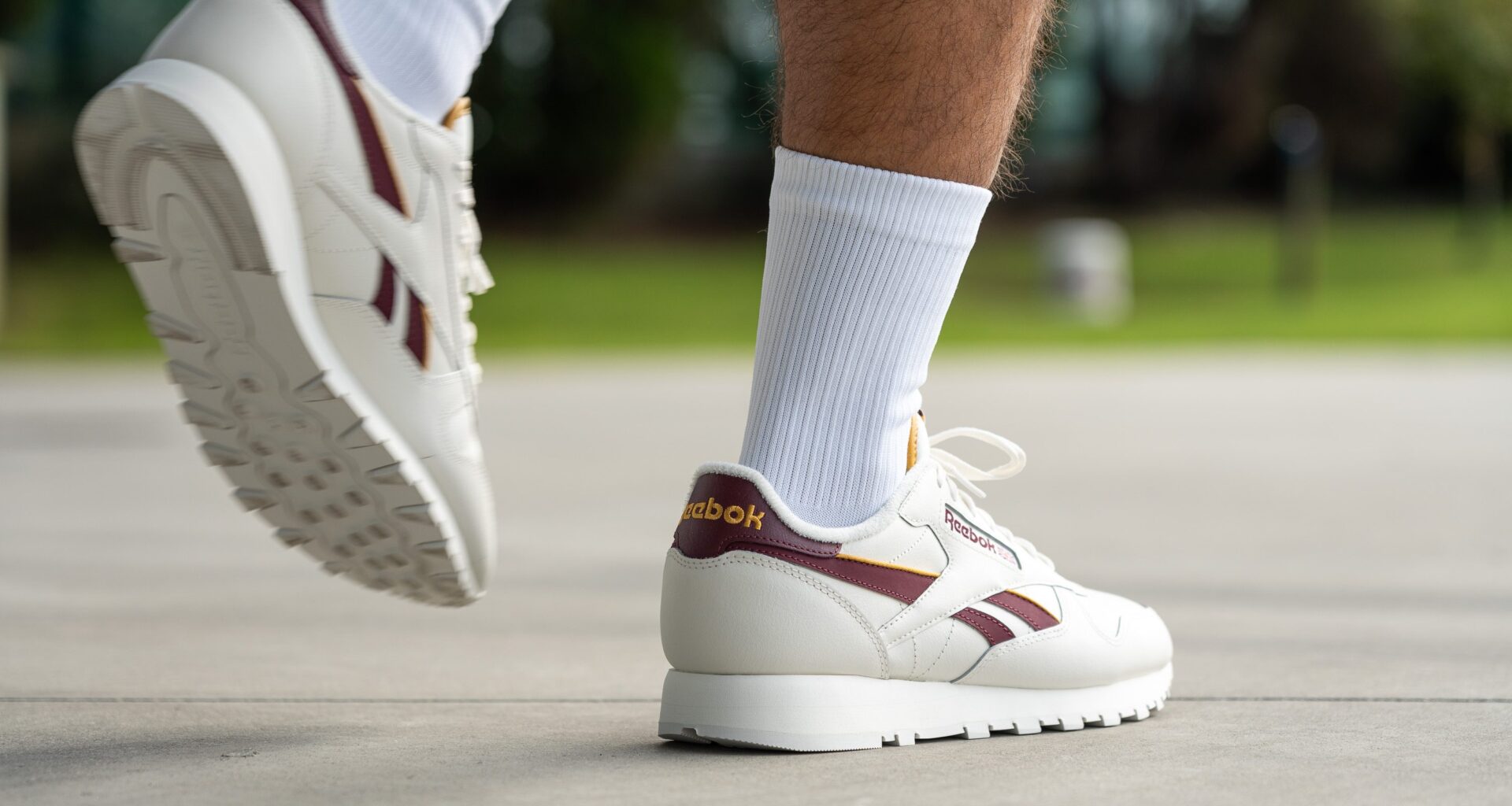 Muž v bílých teniskách Reebok s vínovými detaily a bílých ponožkách kráčí po chodníku.