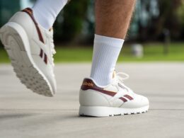 Muž v bílých teniskách Reebok s vínovými detaily a bílých ponožkách kráčí po chodníku.