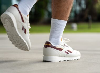 Muž v bílých teniskách Reebok s vínovými detaily a bílých ponožkách kráčí po chodníku.