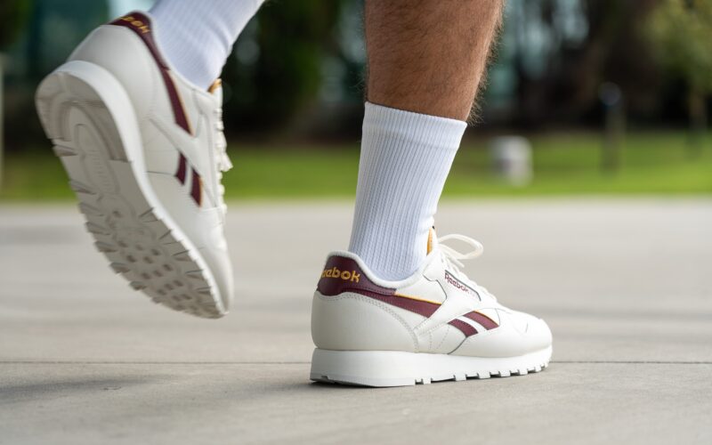 Muž v bílých teniskách Reebok s vínovými detaily a bílých ponožkách kráčí po chodníku.