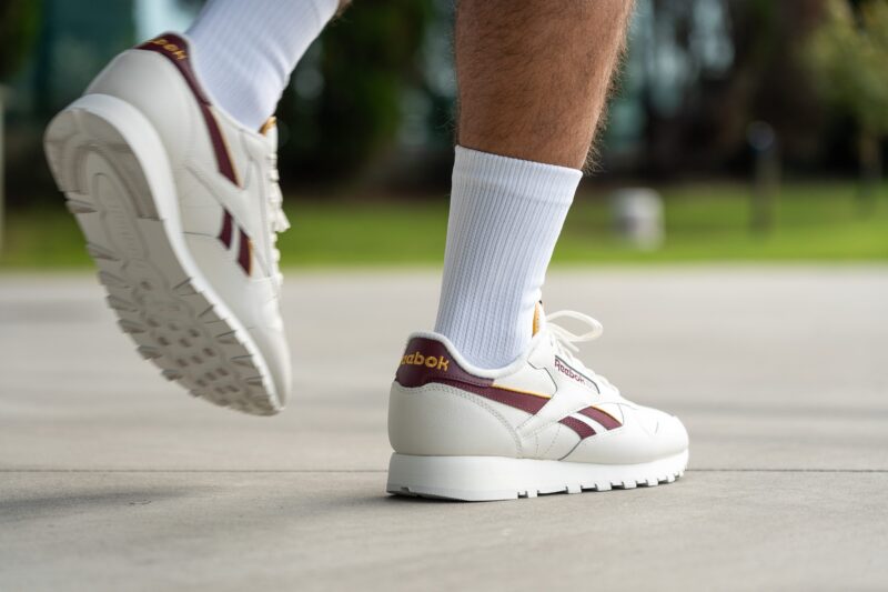 Muž v bílých teniskách Reebok s vínovými detaily a bílých ponožkách kráčí po chodníku.