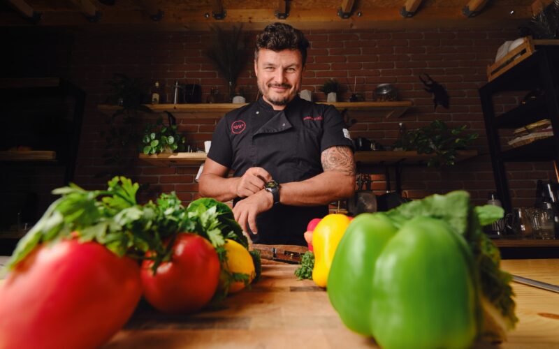 Šéfkuchař stojí za kuchyňským pultem s různobarevnou paprikou a bylinkami v popředí.