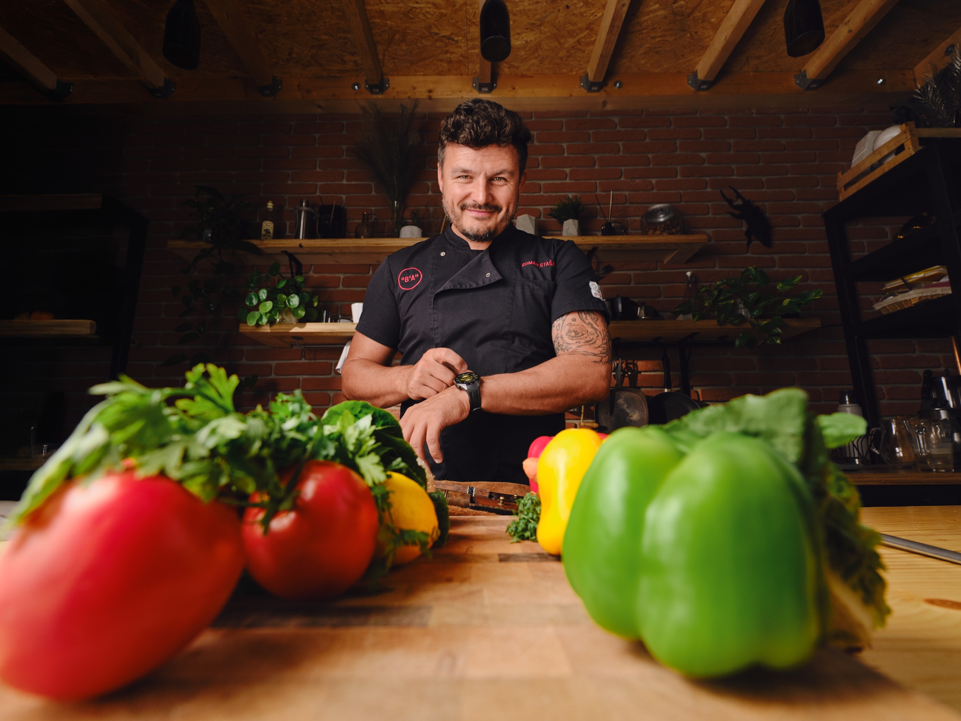 Šéfkuchař stojí za kuchyňským pultem s různobarevnou paprikou a bylinkami v popředí.