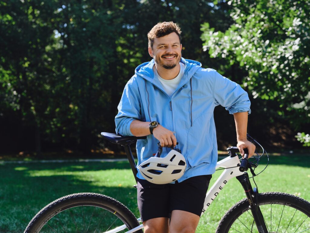 Muž v modré bundě stojí vedle kola, drží cyklistickou přilbu a usmívá se.