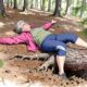 Starší žena ležící na spadlém kmeni stromu v lese s rozpaženýma rukama a zavřenýma očima.