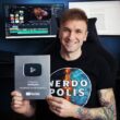 Usměvavý muž v tričku Nerdopolis drží stříbrné YouTube ocenění za 100 000 odběratelů, s tetováním a monitory v pozadí.