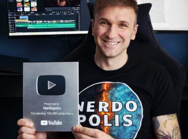Usměvavý muž v tričku Nerdopolis drží stříbrné YouTube ocenění za 100 000 odběratelů, s tetováním a monitory v pozadí.