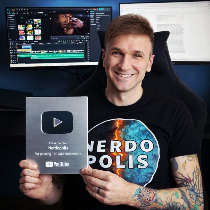 Usměvavý muž v tričku Nerdopolis drží stříbrné YouTube ocenění za 100 000 odběratelů, s tetováním a monitory v pozadí.